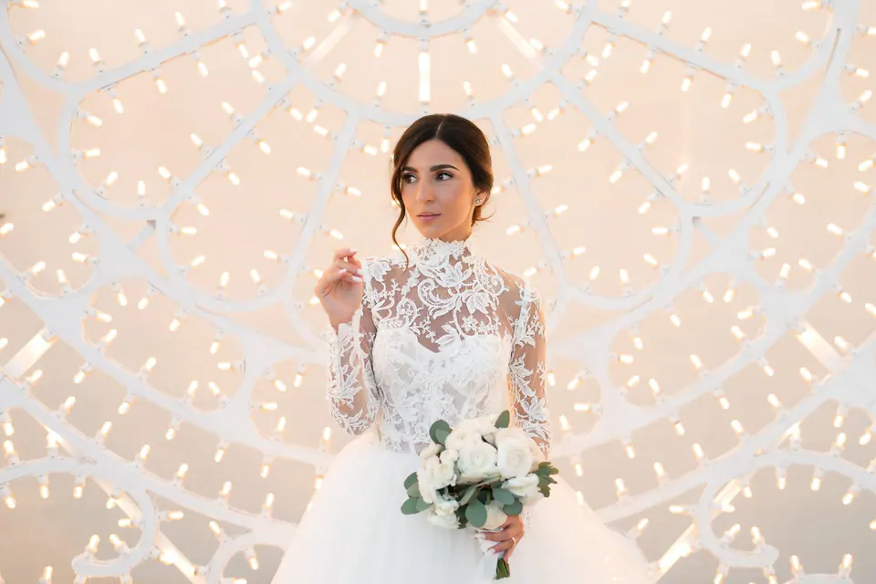 Bride holding her bouquet in front of a wall of small lights in the evening.