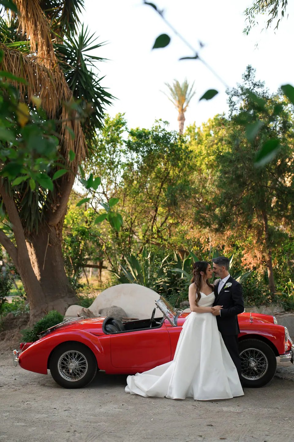 Los novios junto a un descapotable antiguo rojo en el jardín.