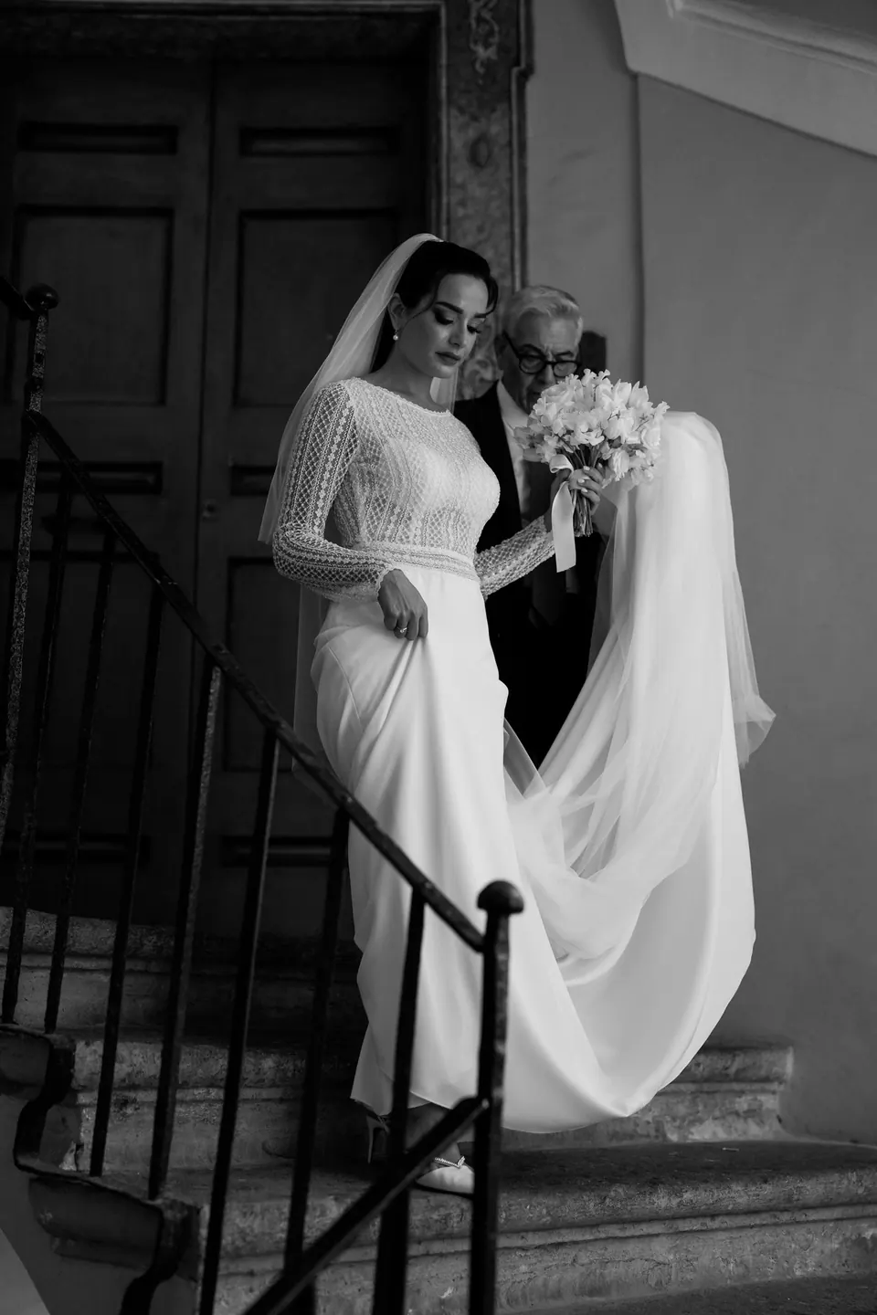 Black and white photo of the bride coming down a dark staircase, veil and bouquet in hand.