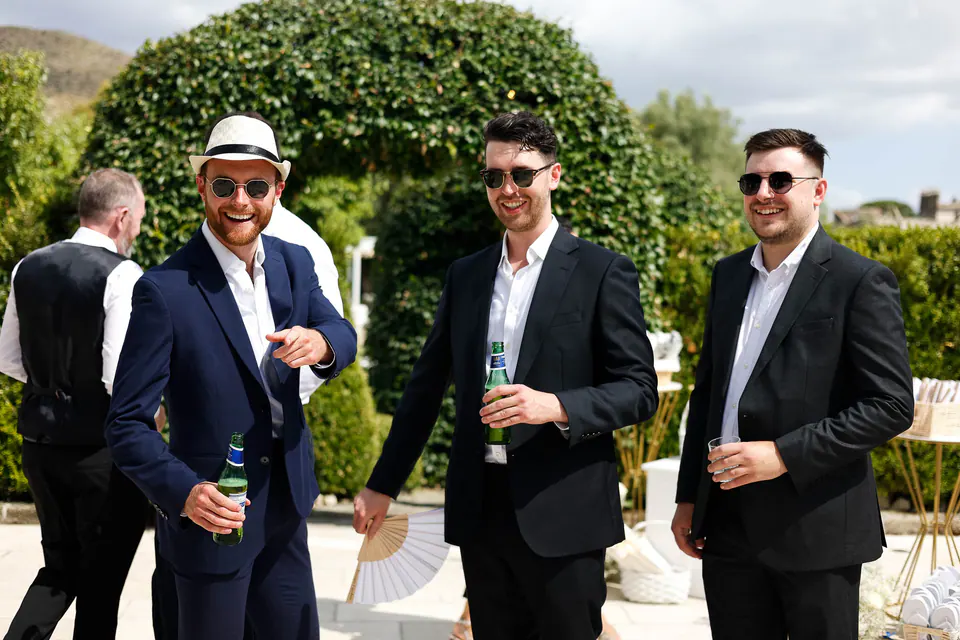 Des invités en costume et un chapeau de paille discutent devant une arche de haie taillée.