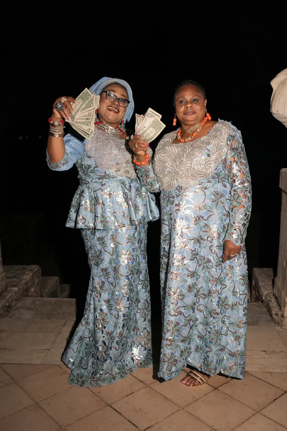 Dos invitados con trajes nigerianos azules y plateados brillantes celebrando por la noche.