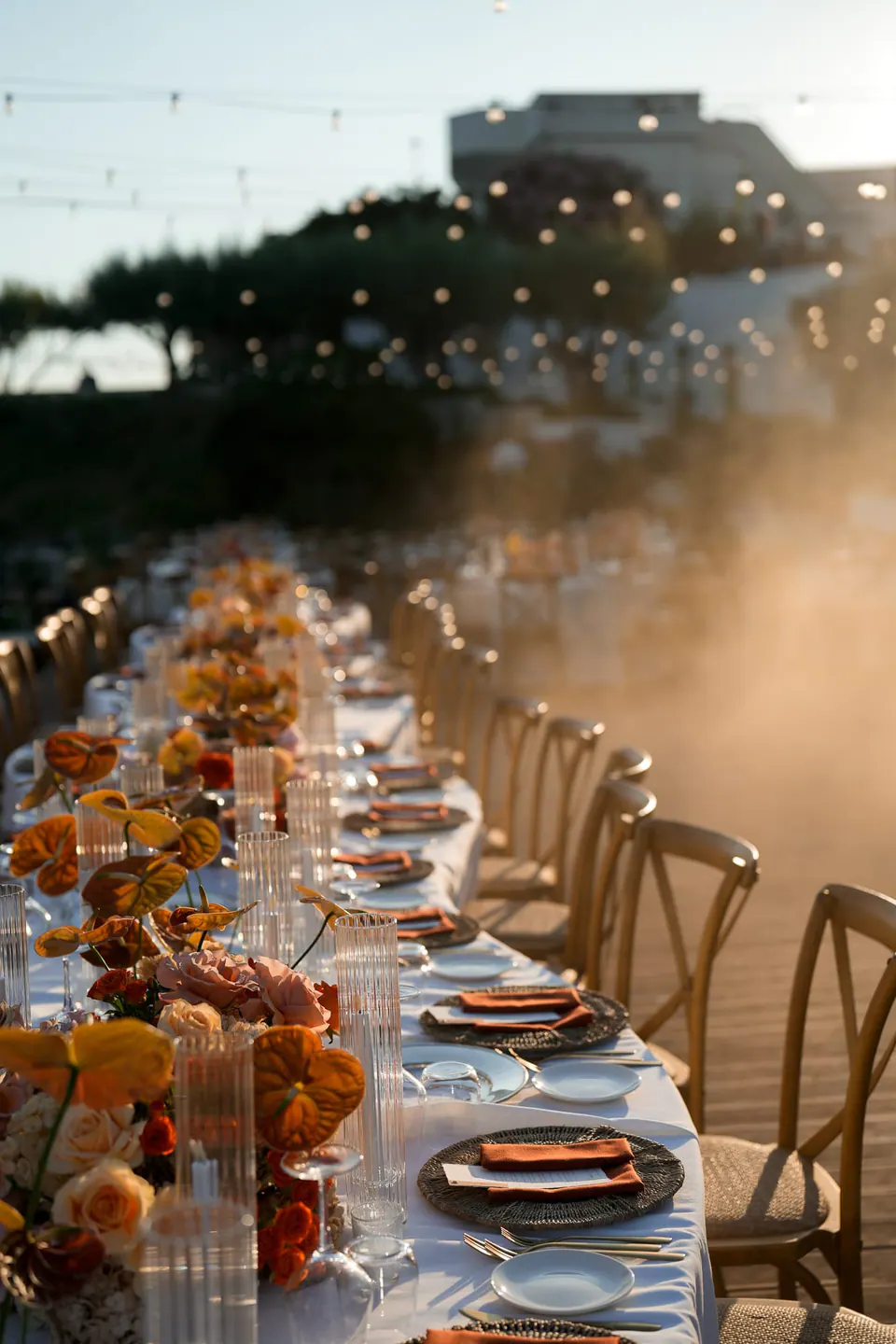 Lange Dinnertafel mit orangen Blumen und Holzstühlen bei Sonnenuntergang über dem Meer.