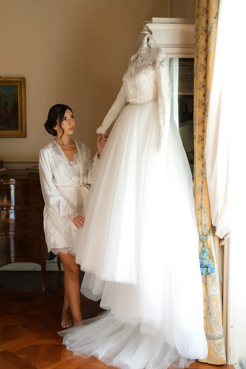 Bride standing next to her wedding dress hanging in a room