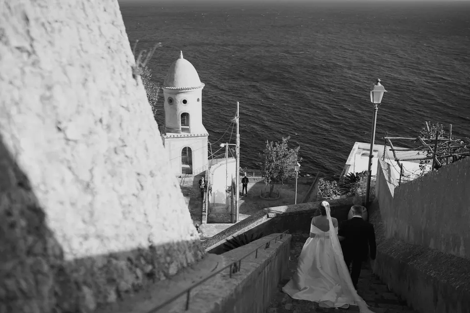 Black and white photo of the couple on a lane beside a bell tower above the sea.