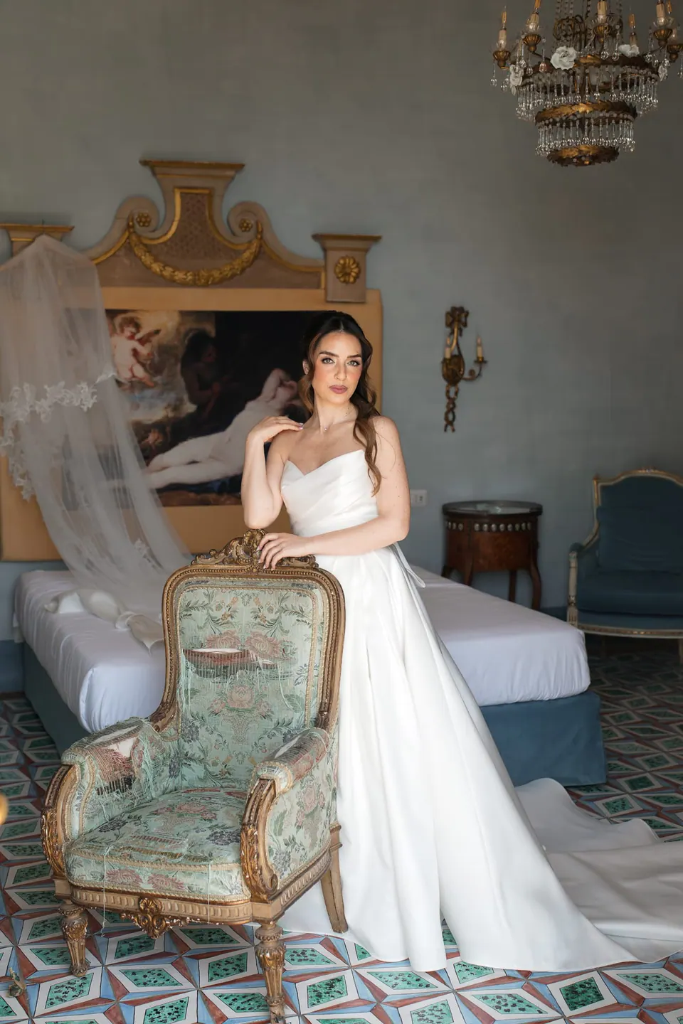 La novia vestida sentada en el brazo de un sillón, con la cama y la lámpara detrás.