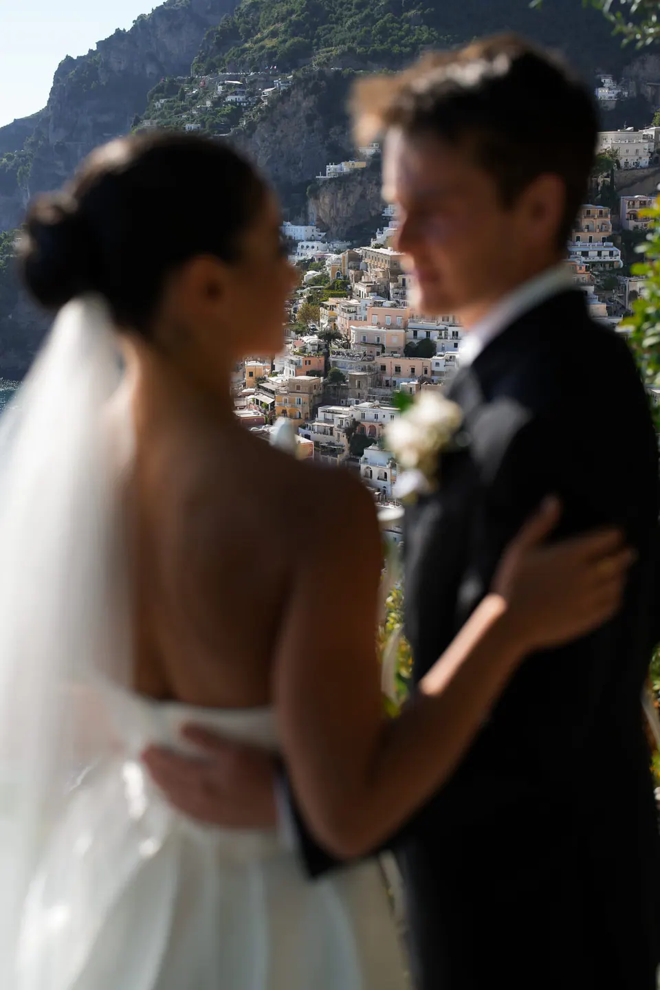 Bride and groom face to face, her veil lit by the sun.