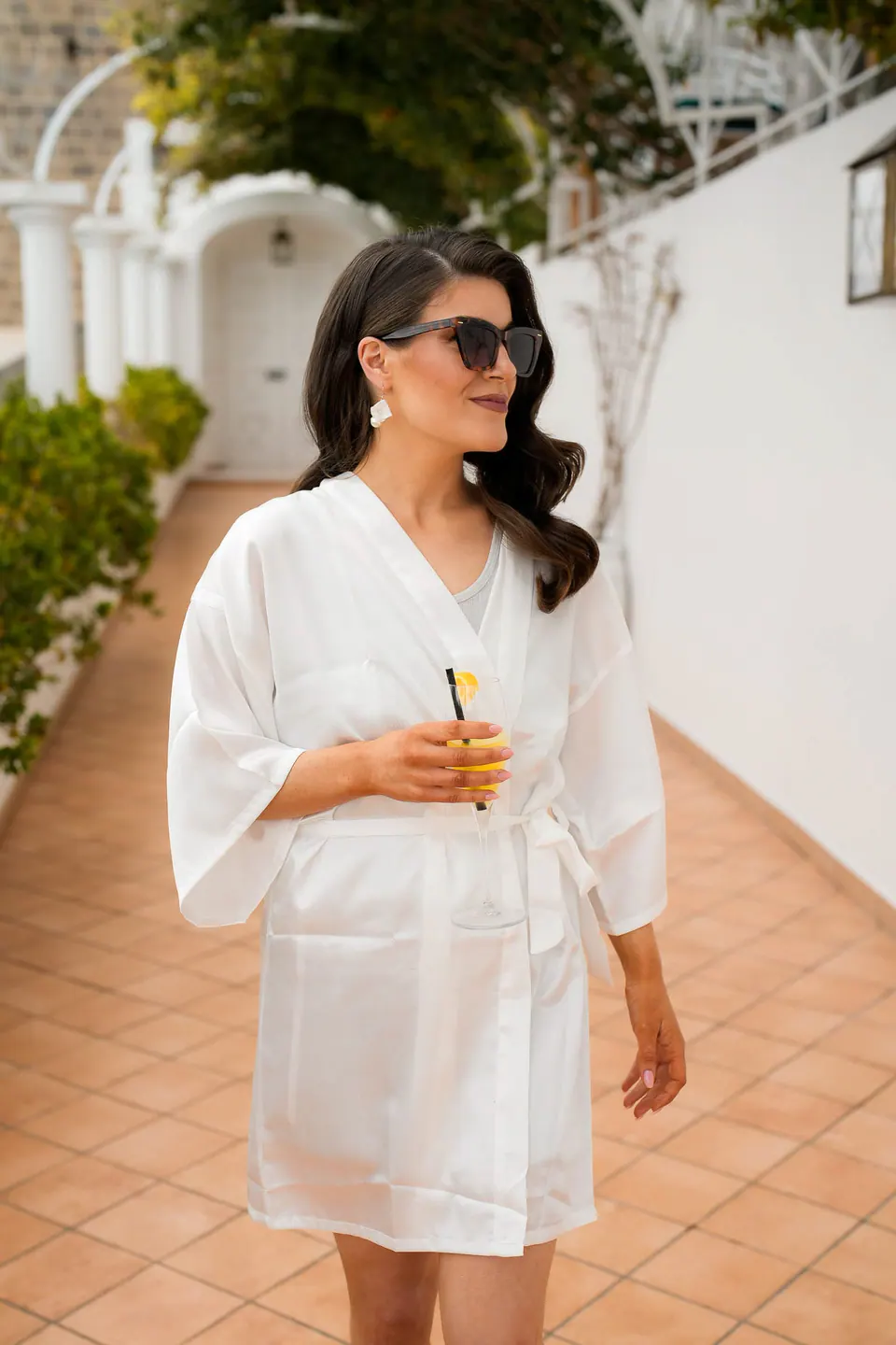 Sposa in vestaglia bianca e occhiali da sole lungo la terrazza della villa, un drink in mano.