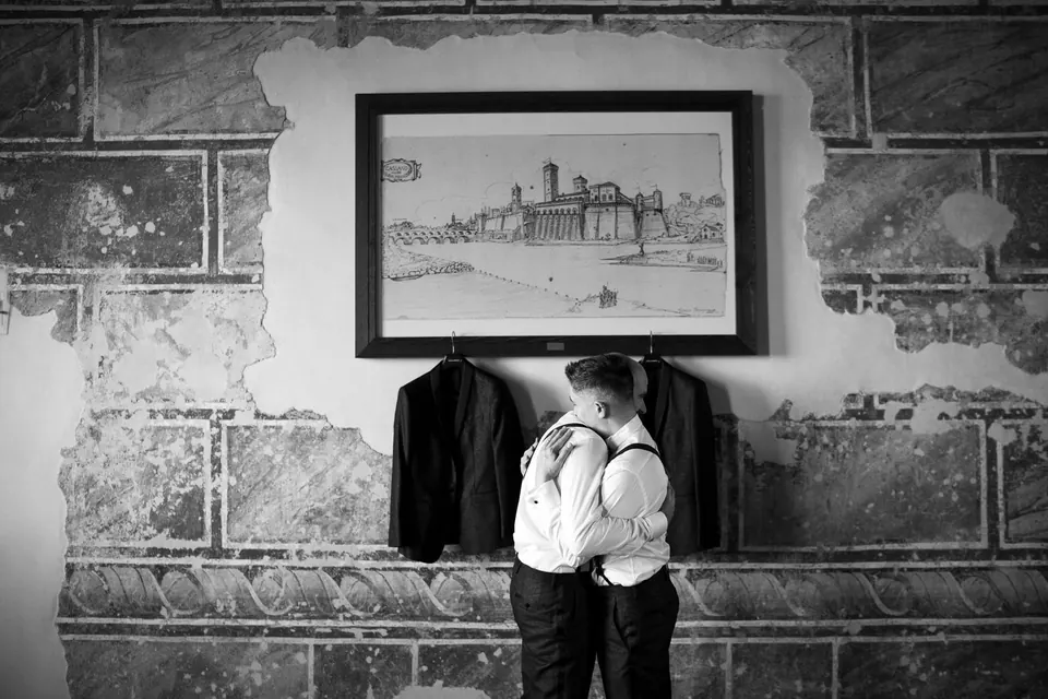 Schwarzweißfoto der Bräutigame in Umarmung unter einem Stich der Burg, die Sakkos an der Wand.