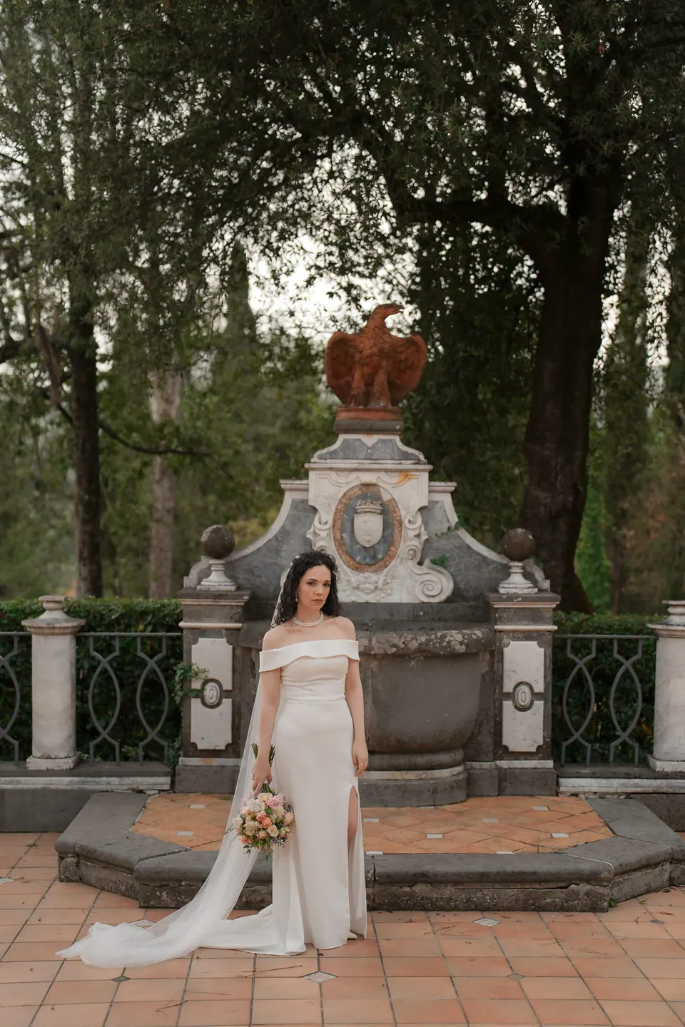 La novia delante de la fuente barroca de la terraza, con el velo extendido sobre las baldosas.