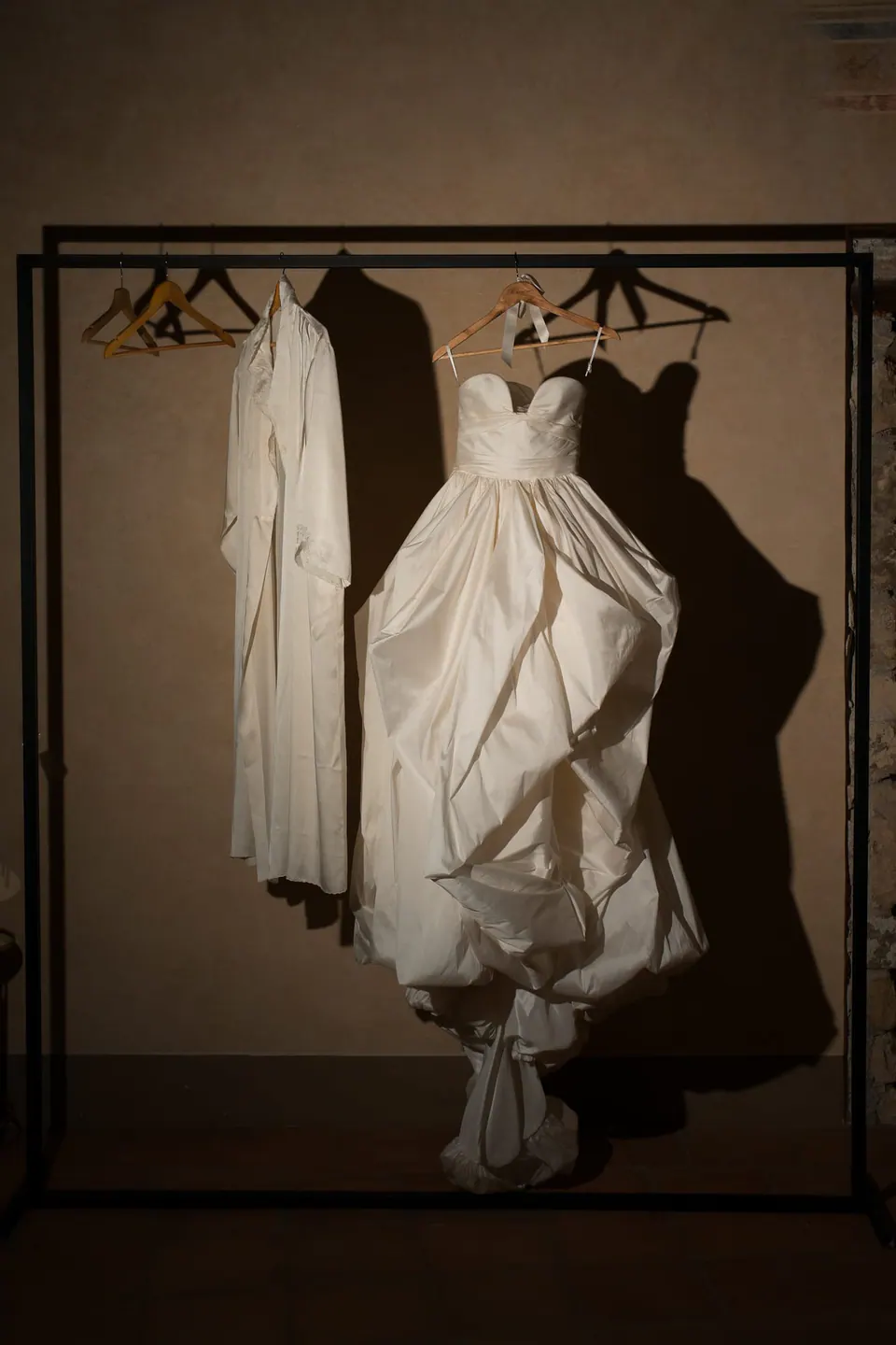 Abito da sposa e vestaglia appesi a uno stender in metallo nero contro una parete calda.