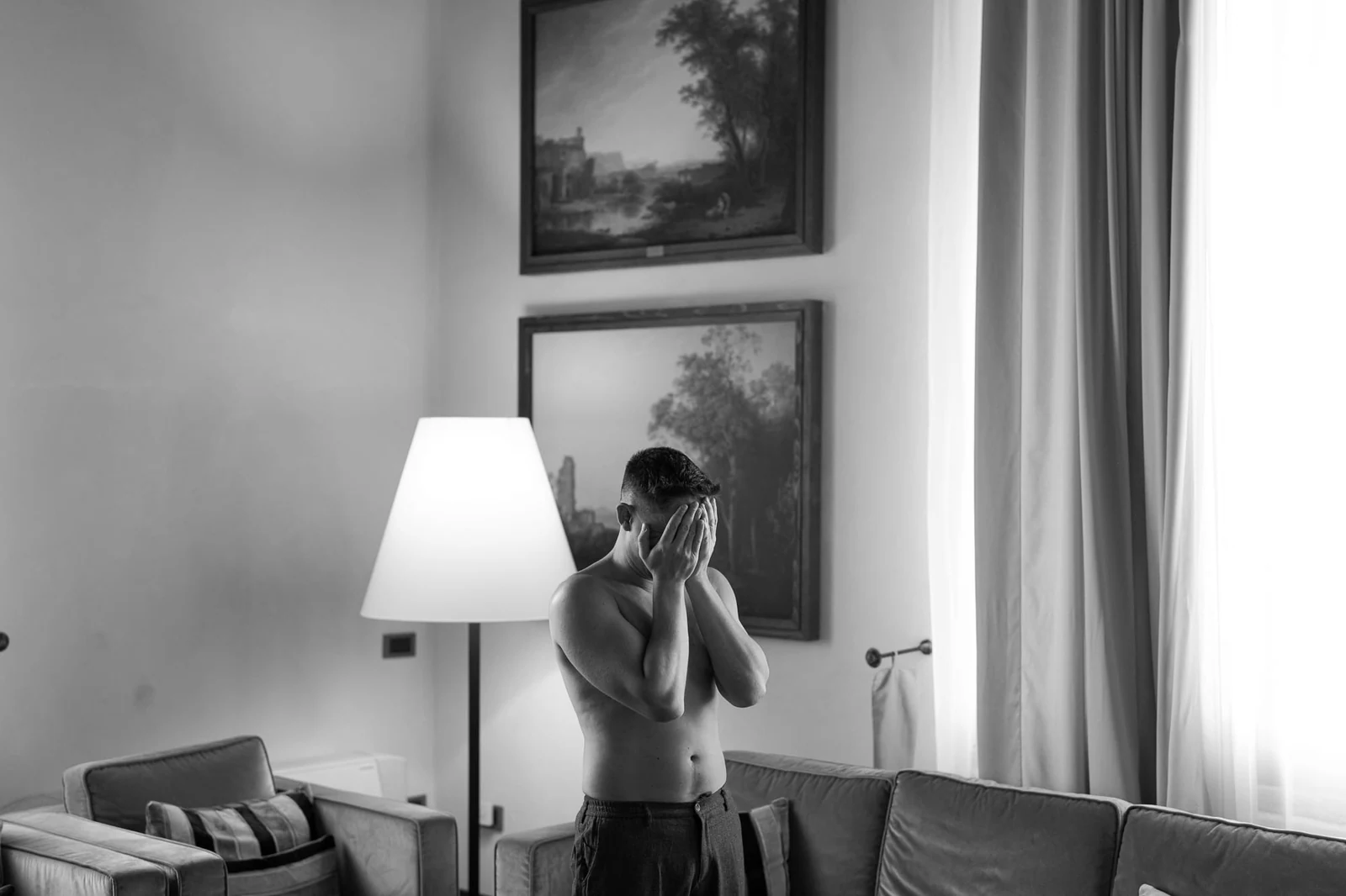 Schwarzweißfoto: Einer der Bräutigame richtet neben einer Lampe in der Suite seine Krawatte.