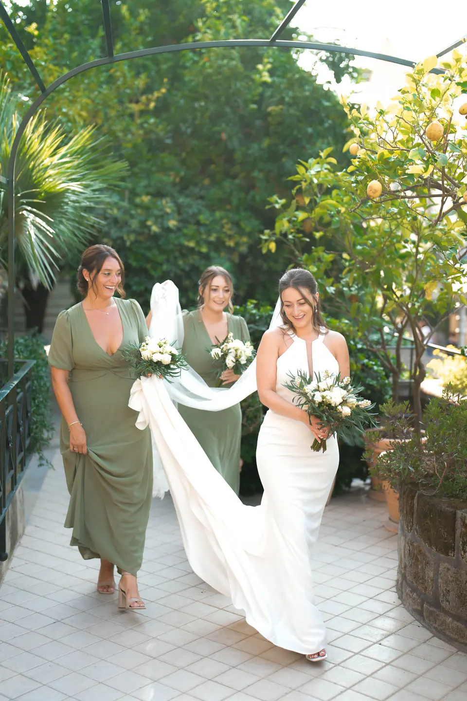 Bride walking to the ceremony at Relais Blu, a bridesmaid in sage green carrying her train.