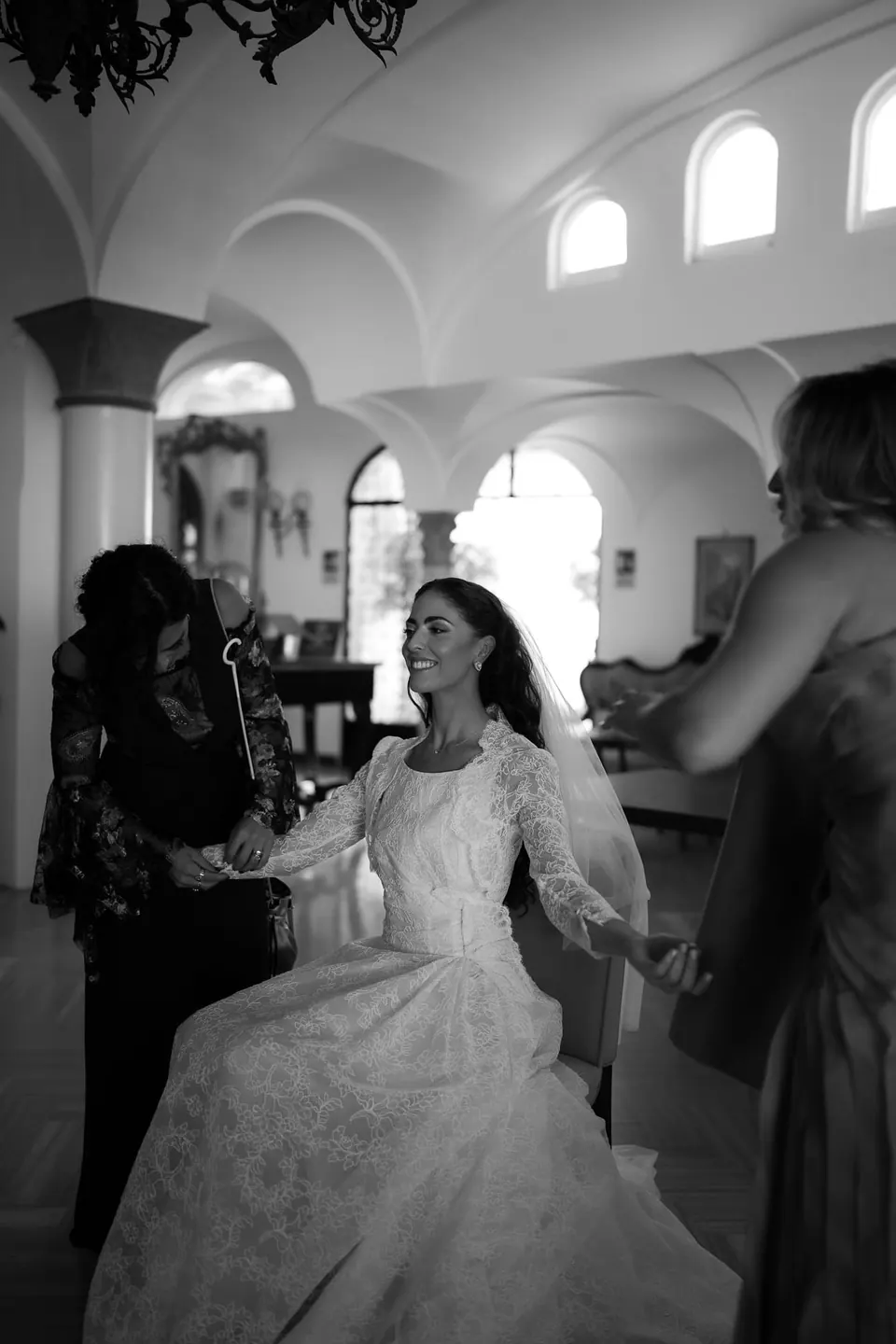 Black and white photo of the bride getting ready among the women of her family in an arched room.