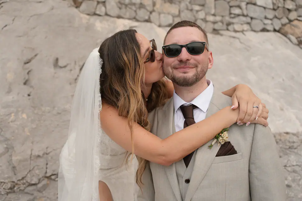 Bride hugging and kissing the groom, both laughing in their sunglasses.