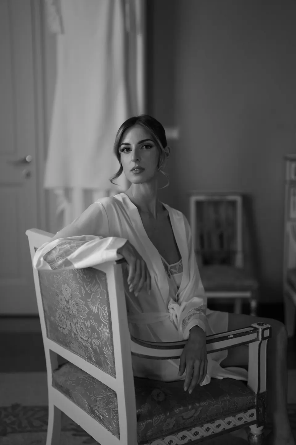 Ritratto in bianco e nero della sposa in vestaglia, seduta di lato su una sedia antica.