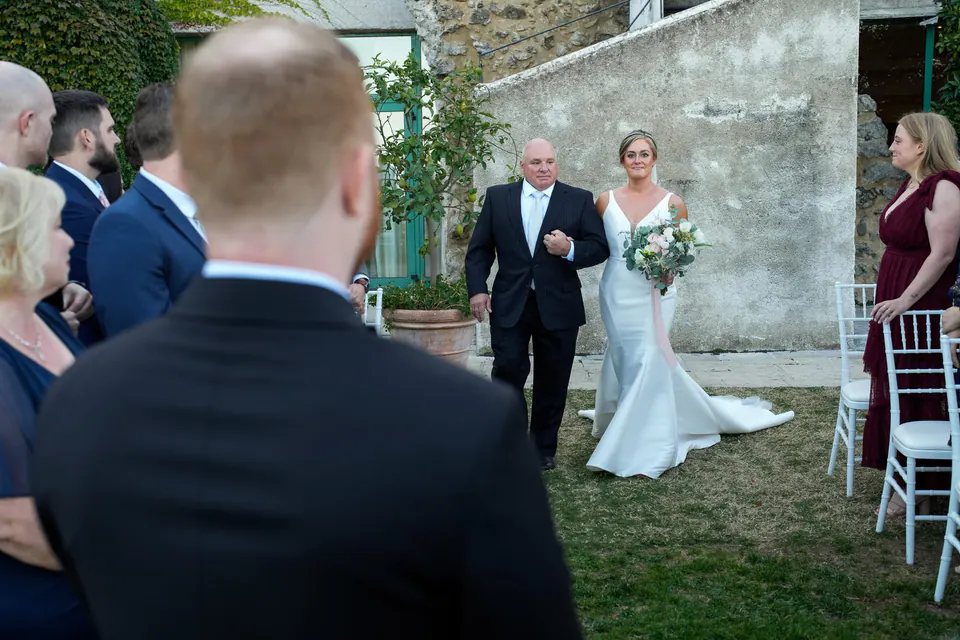 The bride arriving at the ceremony in the garden, the groom watching