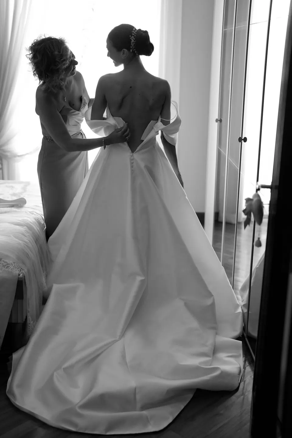 Foto in bianco e nero di una damigella che sistema lo strascico della sposa vicino alla finestra.