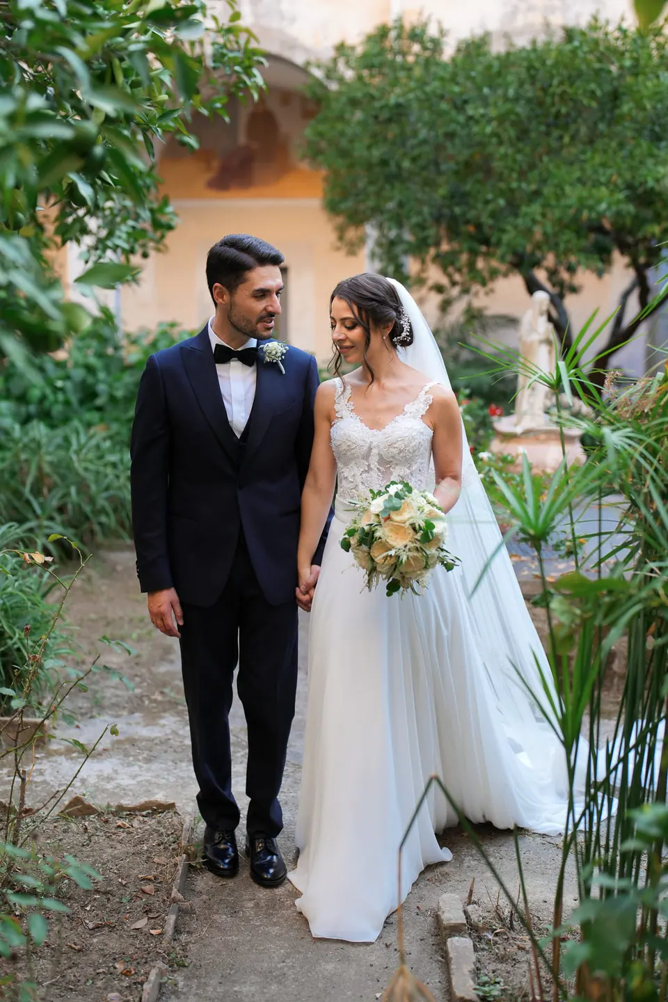 Bride and groom walking down the villa path among the greenery in warm afternoon light.