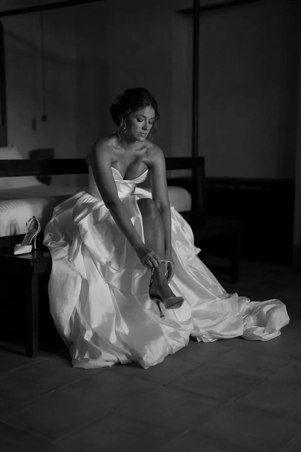 Foto in bianco e nero della sposa seduta sul letto, l'ampia gonna distesa sul pavimento.