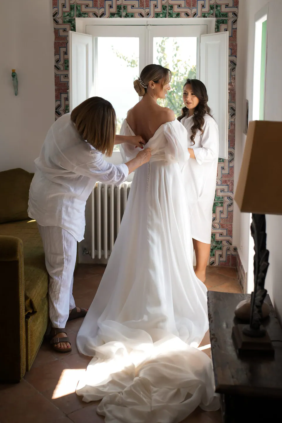 Der Braut wird im Zimmer ins Kleid geholfen, Morgenlicht auf der Schleppe.