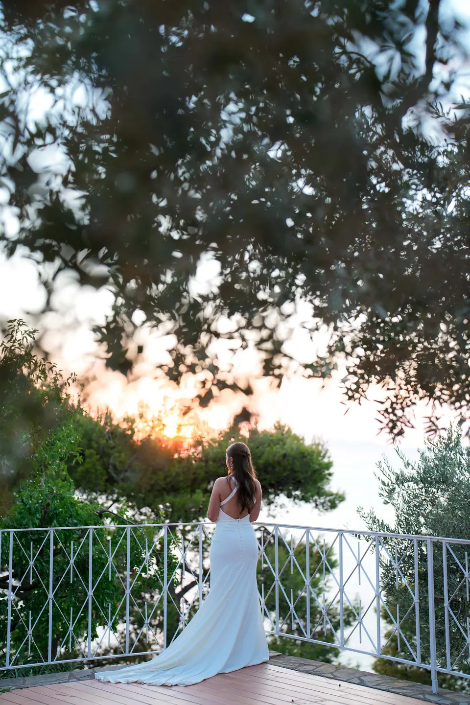 Bride at the balcony railing among the trees, looking out at the sunset.
