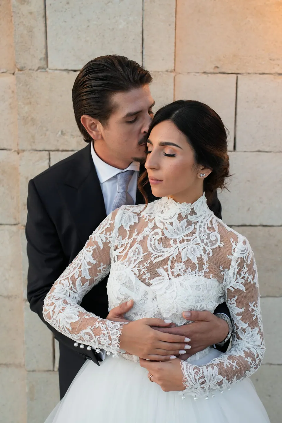 Groom kissing the bride's temple against a stone wall