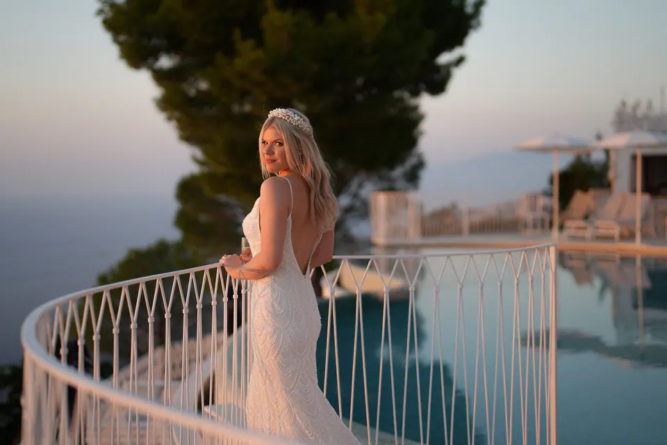 Bride leaning on the terrace railing, veil flowing in the breeze with the sea behind her.