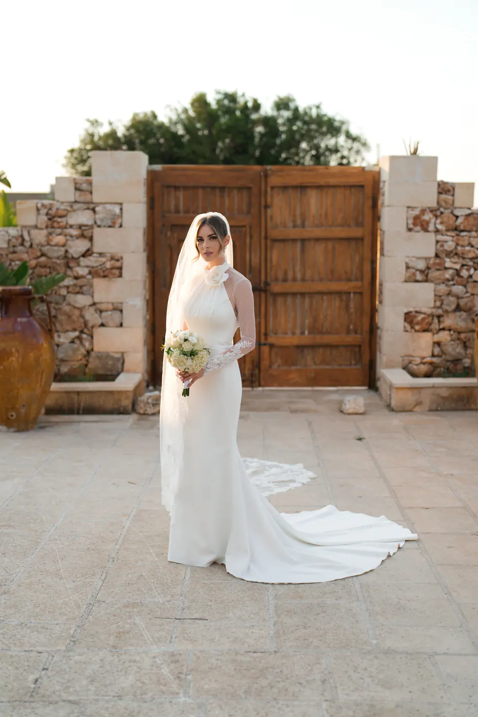 La sposa con il bouquet davanti al portone in legno di Tenuta Sant'Elia al tramonto.