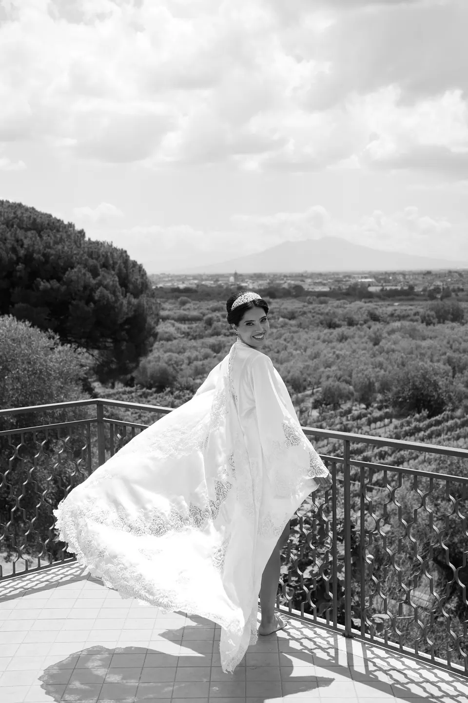 La mariée sur un balcon en noir et blanc, son long peignoir soulevé par le vent au-dessus des oliveraies.