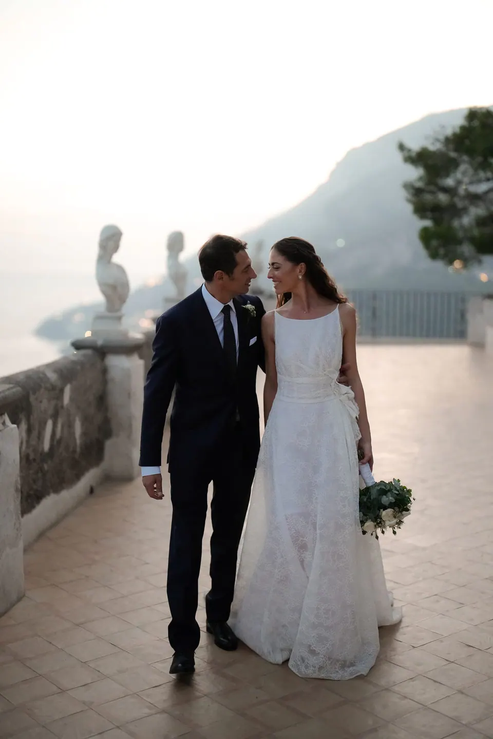 Bride and groom walking on the Terrace of Infinity at Villa Cimbrone, marble busts behind them.