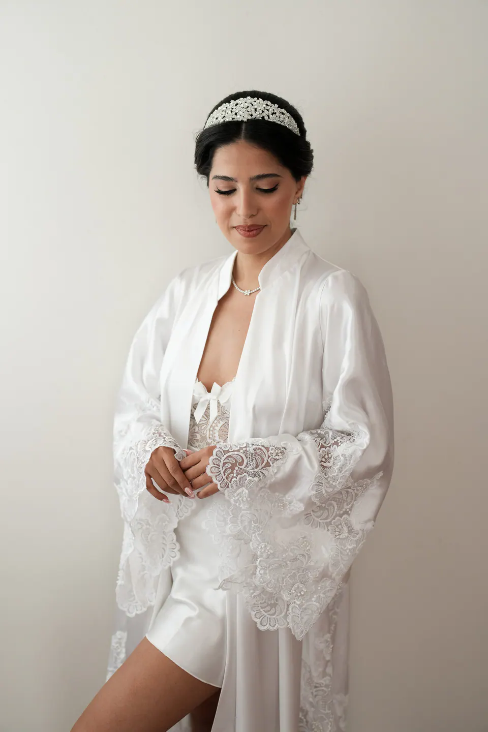 La mariée, le regard baissé, en peignoir et nuisette de satin contre un mur clair.