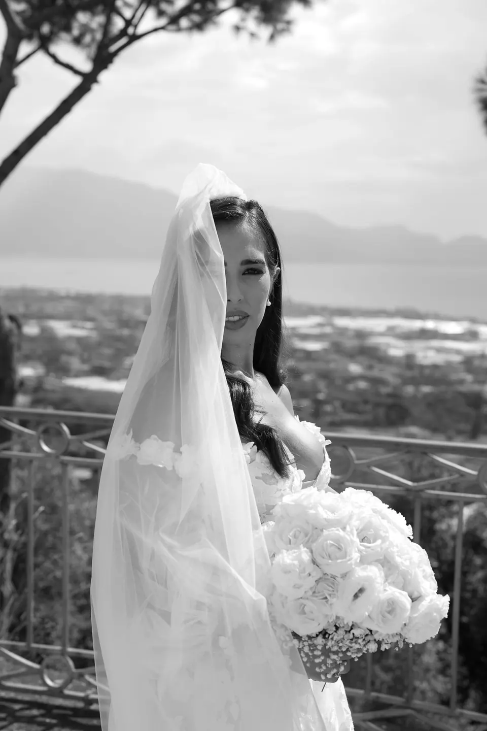 Black and white portrait of the bride in her veil holding white roses, the bay of Pozzuoli below.