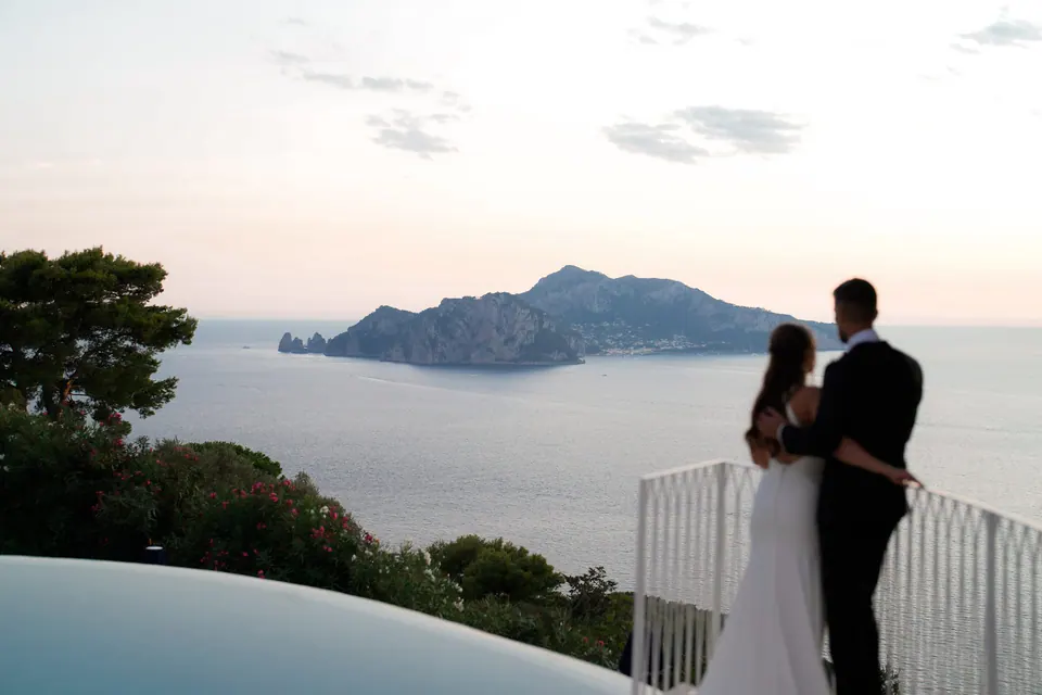 Bride and groom at the pool railing looking toward Capri at sunset.