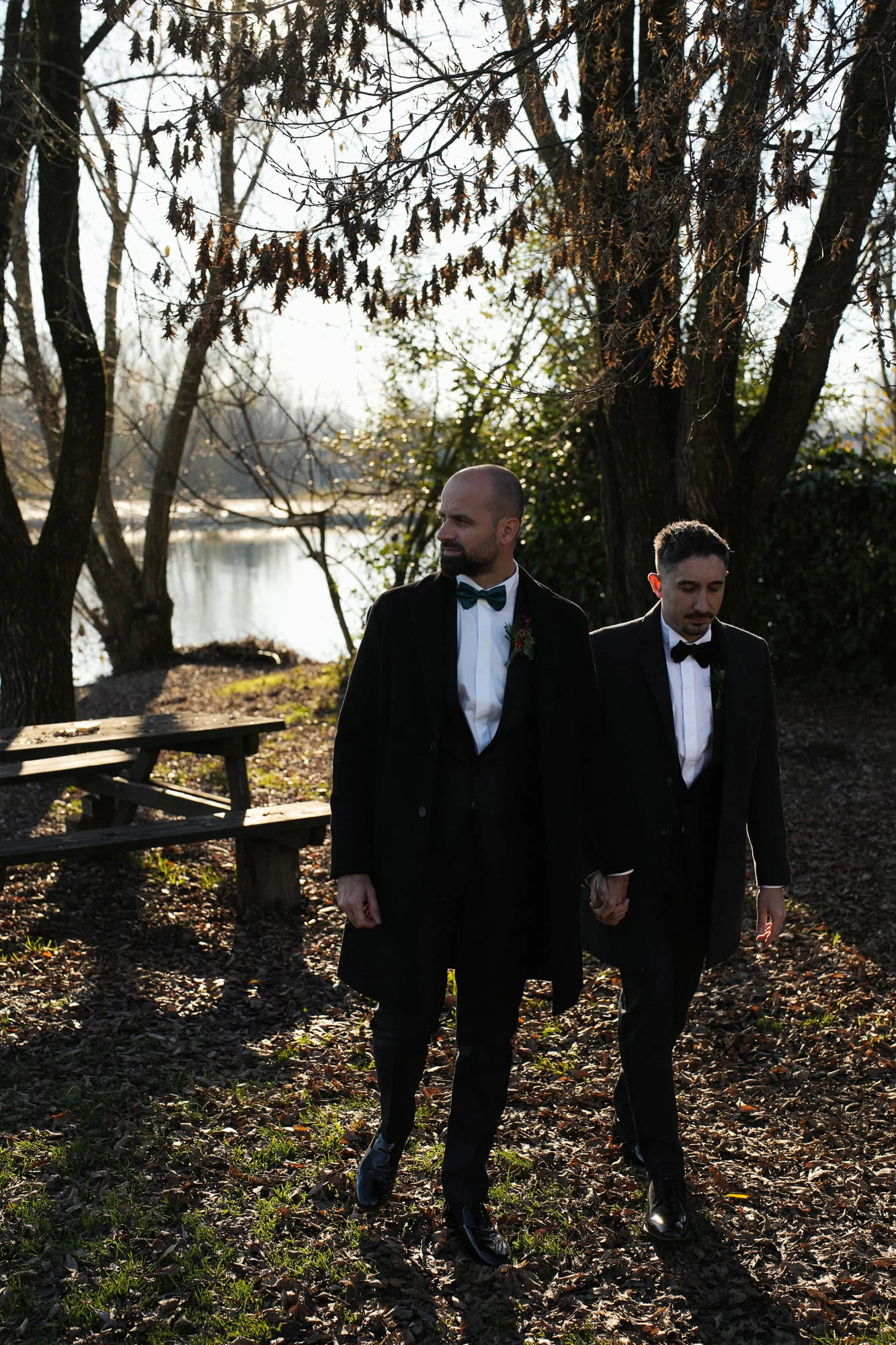 The two grooms walking side by side under the trees by the river at Castello Visconteo.
