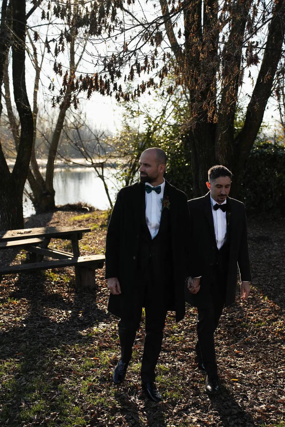 The two grooms walking side by side under the trees by the river at Castello Visconteo.