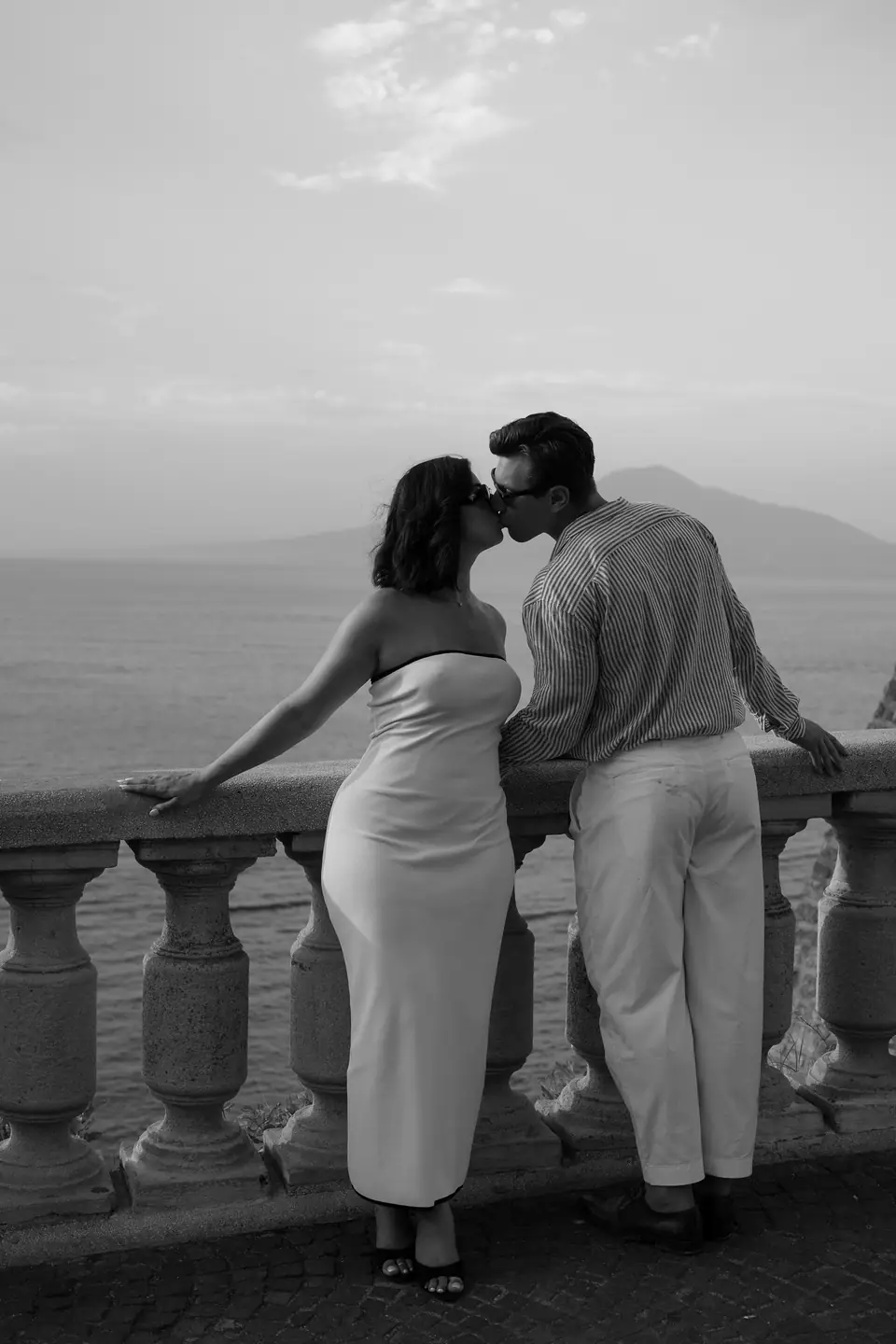 Black and white photo of the couple kissing at a stone balustrade above the bay.