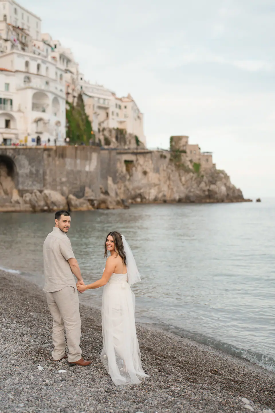 Couple holding hands on the pebble beach, the white houses of Amalfi above them.