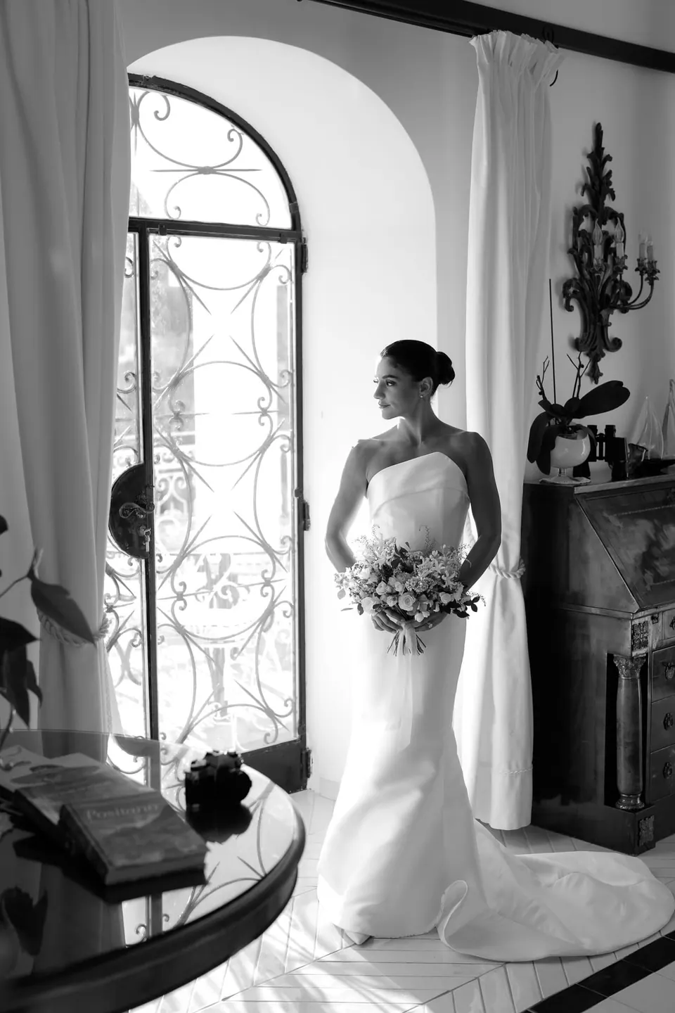 Black and white photo of the bride by an arched window, bouquet in hand.