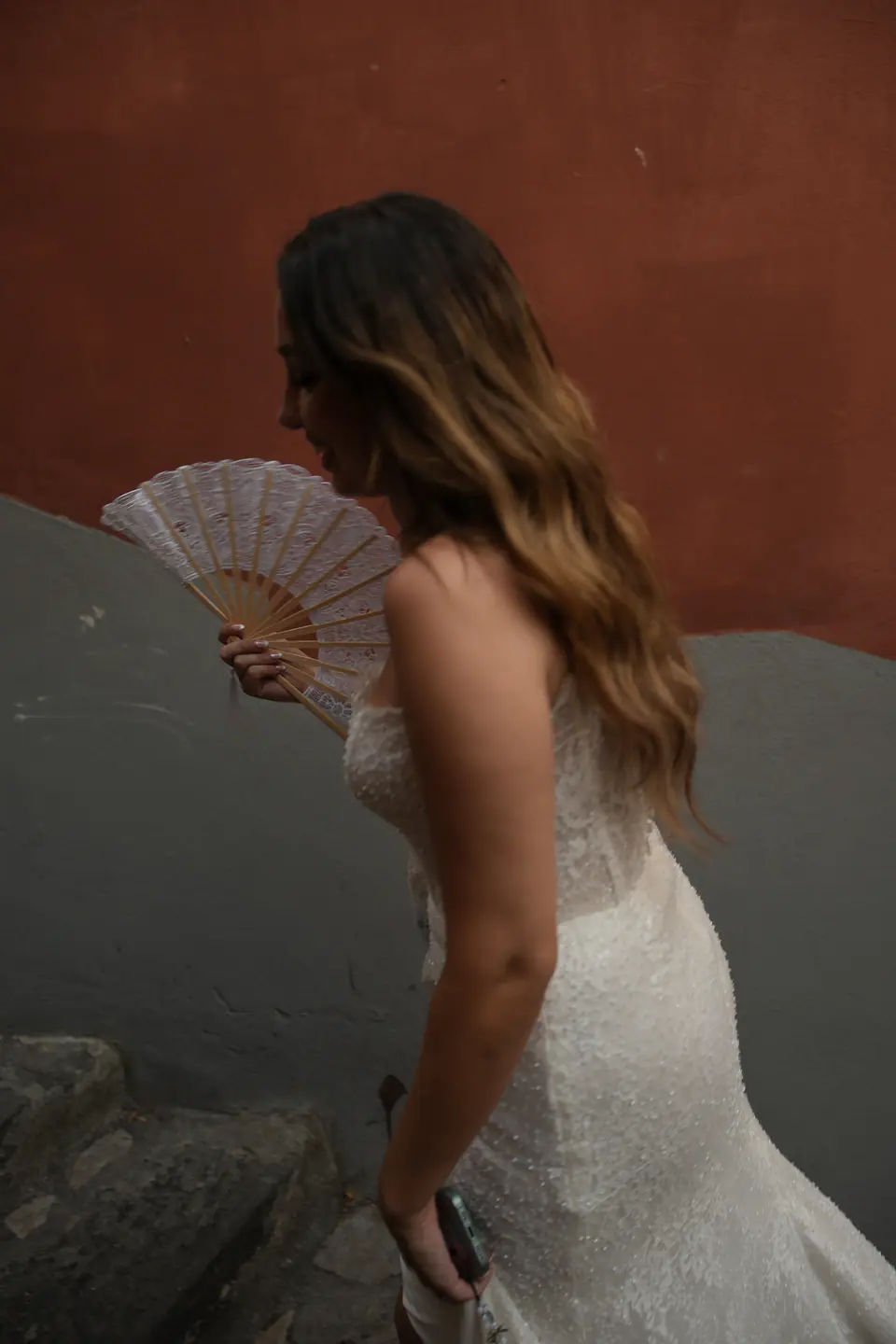 Bride holding a lace fan against a terracotta wall.