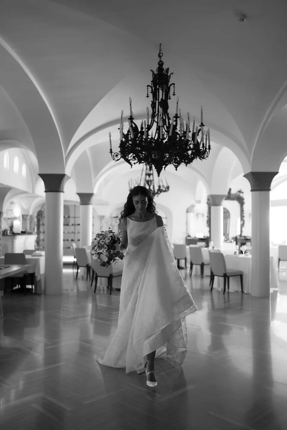 Black and white photo of the bride in her robe in a vaulted hall with a chandelier.