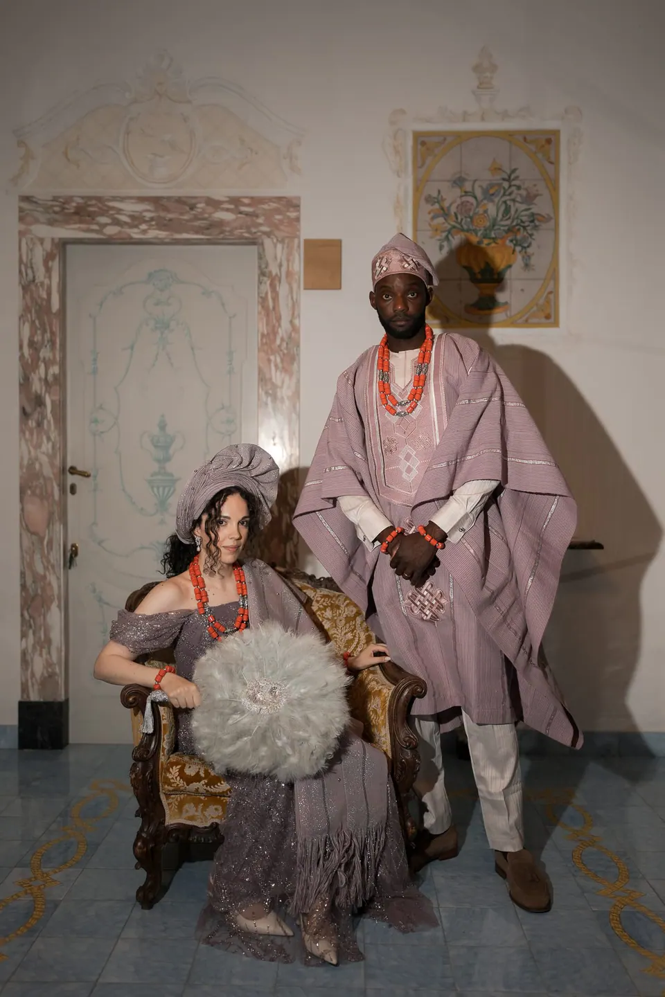Los novios con traje tradicional nigeriano, él con agbada rosa y cuentas de coral, ella sentada con un abanico.