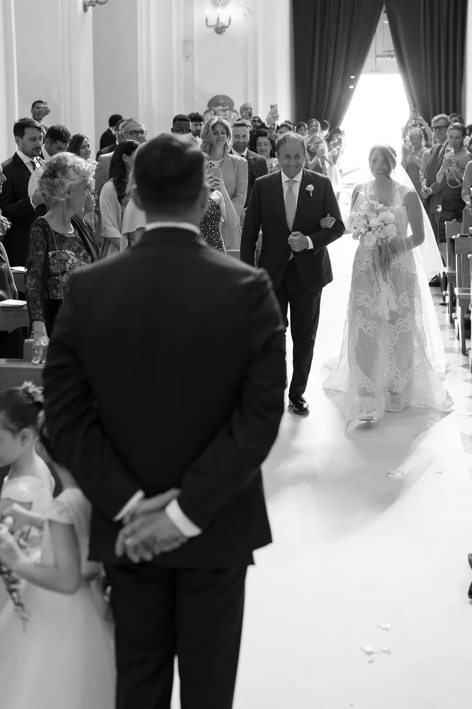 Schwarzweißfoto: Der Bräutigam sieht die Braut mit ihrem Vater durch den Mittelgang kommen.