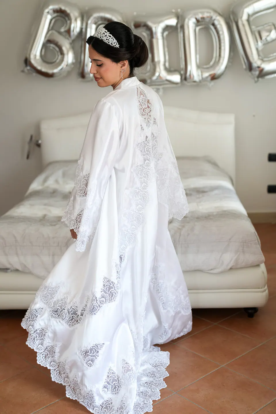 La mariée tourne dans son long peignoir de dentelle devant le lit, des ballons argentés BRIDE au mur.