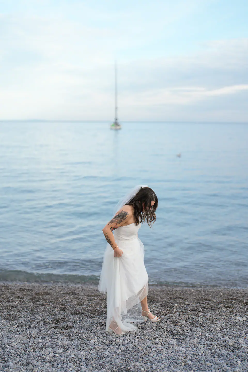 Bride lifting her dress at the water's edge, a sailing boat on the horizon.