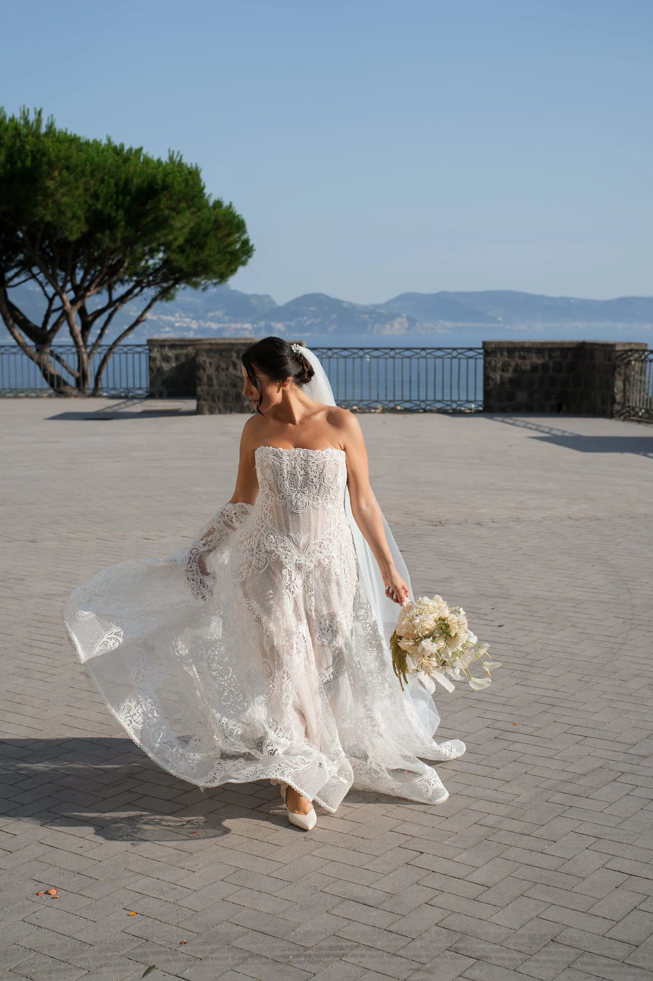 La novia cruza una plaza soleada hacia el mar, con pinos en el horizonte.