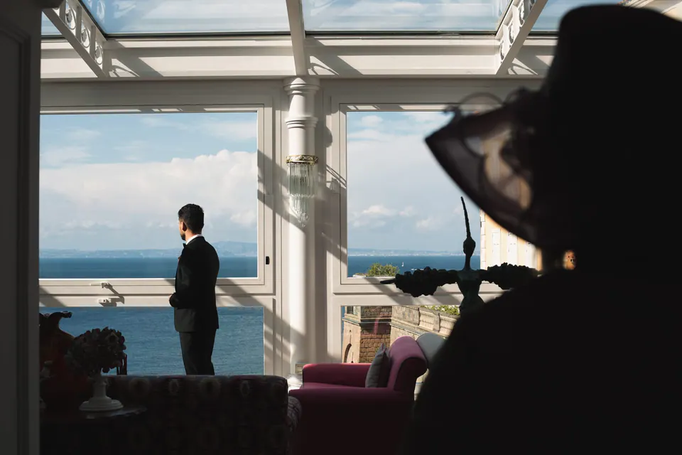 Groom standing at the window before the ceremony, a guest's black hat in the foreground.