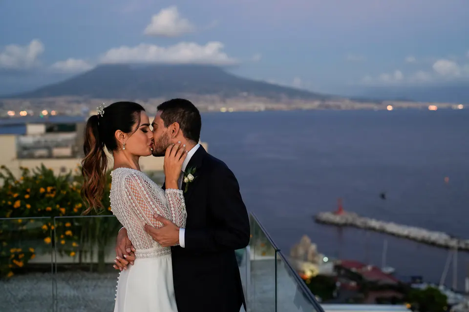 Bride and groom kissing on the terrace of Hotel Vesuvio, Vesuvius and the Gulf of Naples behind them.