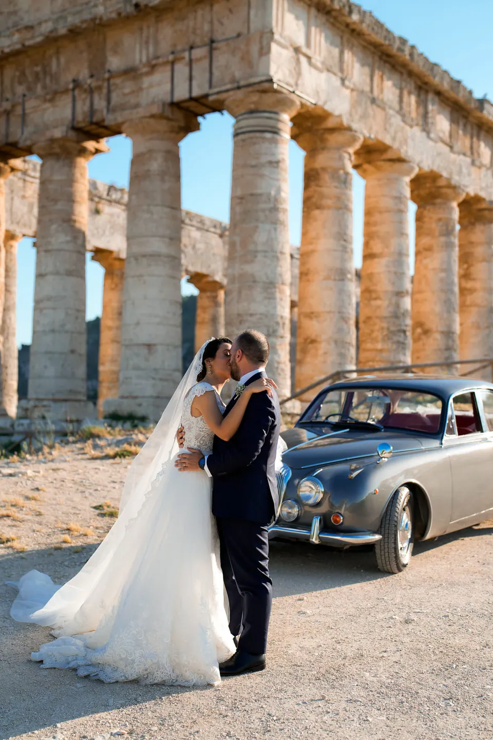 Bride and groom kissing in front of an ancient Greek temple in Sicily, a vintage car beside them