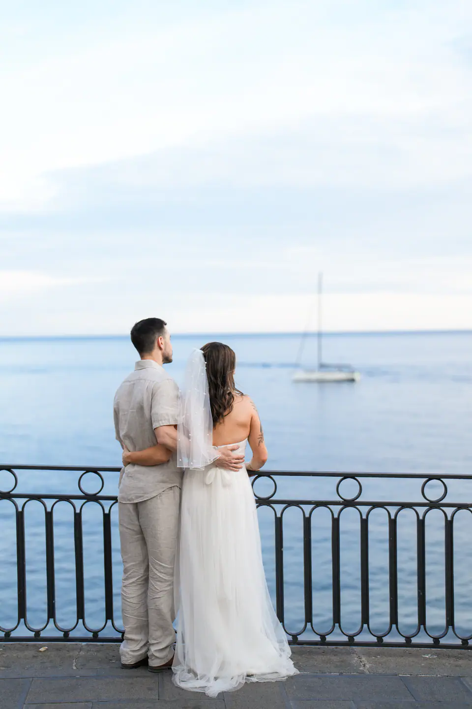 Couple seen from behind at a railing, looking out to the sea and a sailing boat.