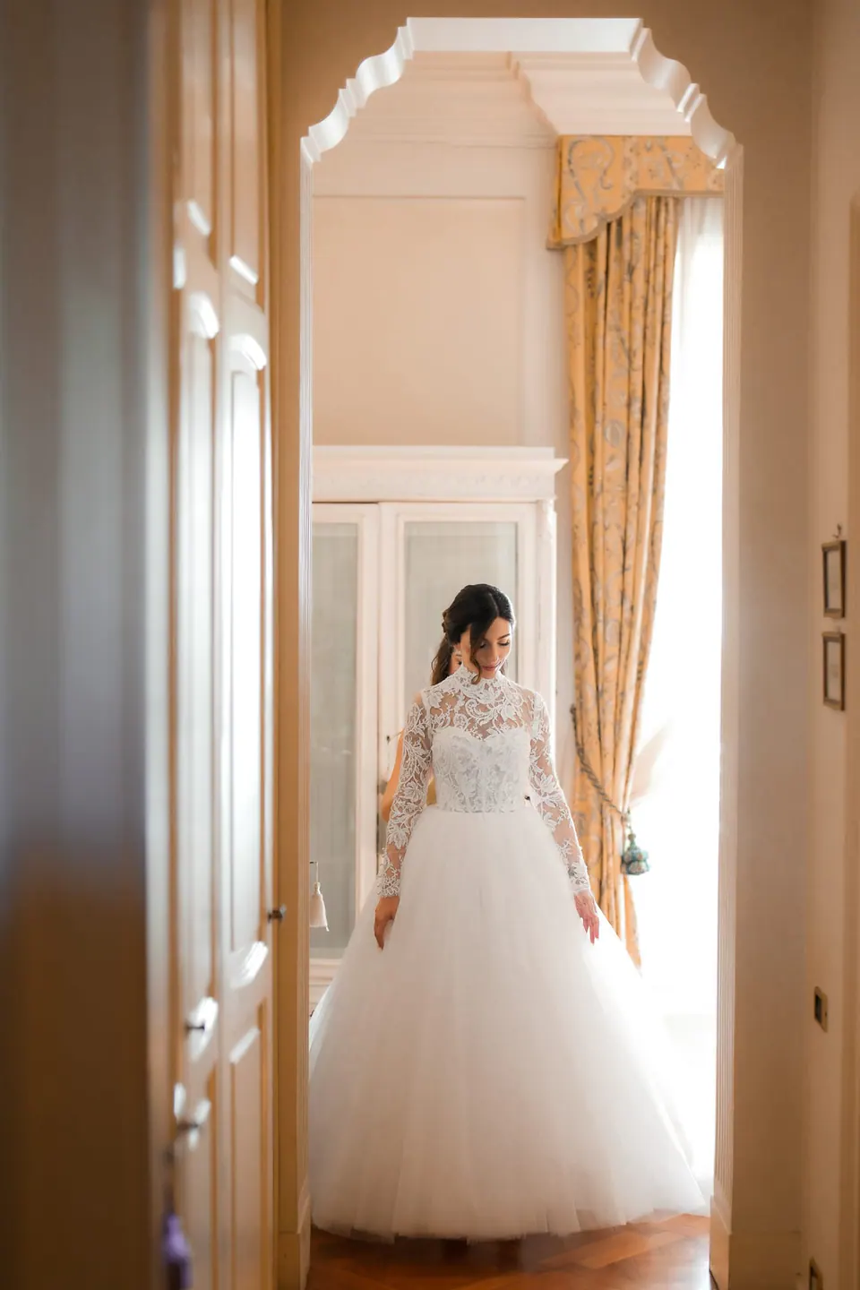 Bride standing gracefully by a window, veil and gown flowing softly in the golden hour light.